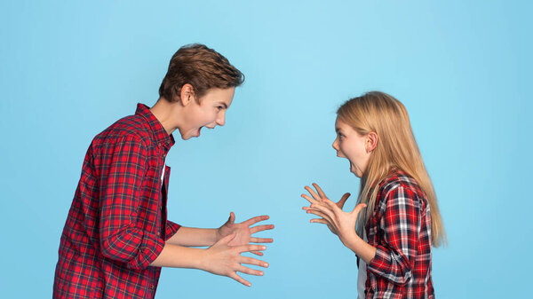 Siblings Conflicts. Angry Little Brother And Sister Emotionally Screaming At Each Other, Standing On Blue Background With Free Space