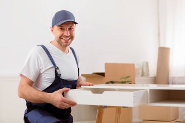 Tamirci, mobilyaları boşaltıyor, mobilyaları yerleştiriyor, mobilyaları yerleştirdikten sonra evde çalışıyor, kameraya gülümsüyor.