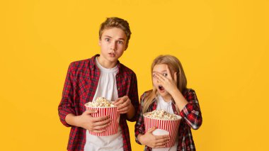 Scary movie. Frightened kids brother and sister holding popcorn buckets and looking at camera, little girl peeking through fingers