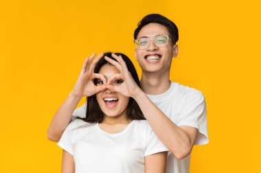 Mutlu Asyalı çift eğleniyor, parmaklarını kız arkadaşının gözlerine yaklaştırıyor, sarı stüdyo geçmişi var, her ikisi de fotokopi odasına bakıyor.