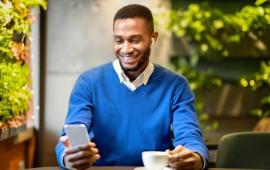 Teknoloji Konsepti. Modern kafede oturan akıllı telefon kullanan kulaklıklı mutlu afro öğrencinin ön görüntüsü.