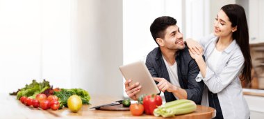 Yeni Tarifler Konsepti. Gülümseyen genç bir aile dijital tablet kullanıyor, mutfaklarında birlikte yemek pişiriyorlar.