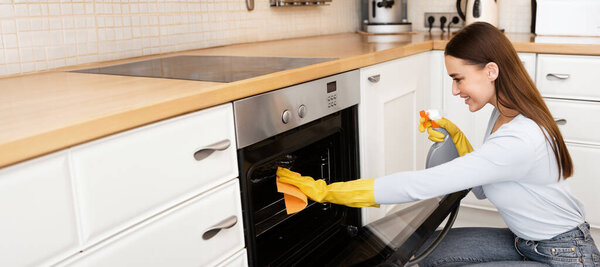 Концепция кухонного дела. Девушка в жёлтых защитных резиновых перчатках моет грязную духовку тряпкой из микроволокна и спреем
