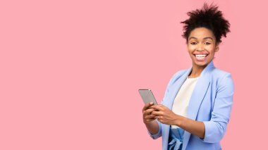 Chatting With Friends. Happy black woman holding mobile phone, looking at camera over pink studio wall, free space