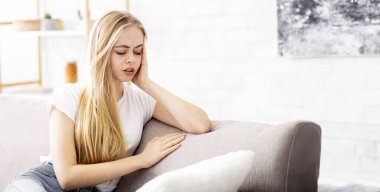 Blonde upset girl sitting on sofa at home, suffering from migrane