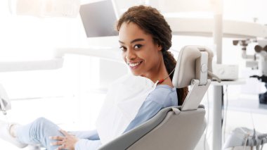 Dişçi koltuğunda oturan ve kameraya gülümseyen neşeli Afrikalı kadının yan görüntüsü.