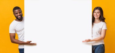 Gülümseyen, çok ırklı bir çift, beyaz reklam kurulunu işaret ediyor, sarı arka planda yan yana duruyorlar, mesajlarınız için bedava bir yer.