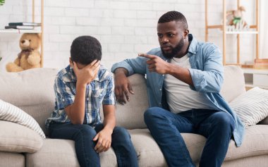 Kızgın siyah baba, evdeki kötü davranışları yüzünden inatçı oğlunu azarlıyor, çocuk facepalm hareketi yapıyor, boş alan yaratıyor.