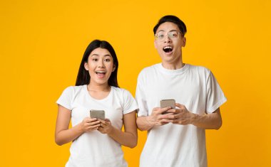 Yeni havalı uygulama. Heyecanlı Asyalı çift, sarı arka planda modern akıllı telefonları tutuyor.