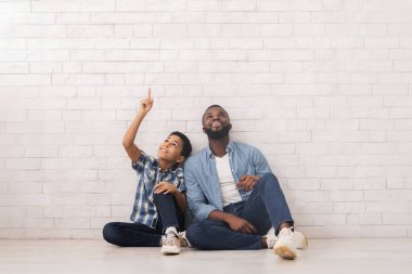 Baba ve oğul beyaz tuğla duvarın yanında oturmuş fotokopi alanını işaret ediyor ve yukarı bakıyor.