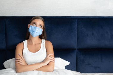 Kendini izole etme. Koruyucu Maskeli Üzgün Kız Karantina Yaşam tarzı ve Yalnızlıktan Sıkılmış Yatakta Oturuyor