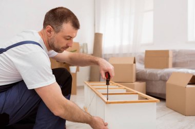 Profesyonel Mobilya Toplayıcı Adam, Odanın zeminine yerleştirildikten sonra ahşap rafı tamir edip monte ediyor. Marangoz 'un Tornavida Aracı Kullanarak Çalışmasına Yan Bakış. Marangozluk Hizmeti Konsepti