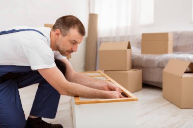 Mavili Kapaklı Tamirci, içeride tadilat sırasında tornavidayla ahşap rafı tamir edip monte ediyor. Mobilya montajı ve marangozluk hizmeti konsepti. Yan Görünüm Görüntüsü