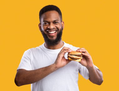Elinde hamburgerle kameraya gülümseyen mutlu siyah adam sarı arka planda duruyor. Stüdyo çekimi. Abur cubur ve aldatma yemeği, beslenme ve erkek diyeti konsepti. Aşırı yeme alışkanlığı