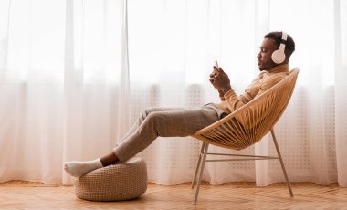Hafta sonu. Kablosuz kulaklıklı gevşek siyah adam cep telefonu kullanıyor Audiobook 'u dinliyor Pencere karşısında modern sandalyede oturuyor