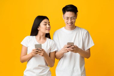 Meraklı Asyalı kız erkek arkadaşını gözetlerken cep telefonu kullanıyor, sarı arka plan.