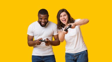 Neşeli ırklar arası çift ellerinde joysticklerle video oyunlarında birbirleriyle yarışıyor, sarı arka planda yan yana duruyorlar, boş alan.