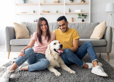 Evcil köpekleri evde yerde oturan neşeli, çok ırklı bir çiftin tam boy portresi. Arap adam ve beyaz karısı Golden Retriever 'la oturma odasında vakit geçiriyorlar.