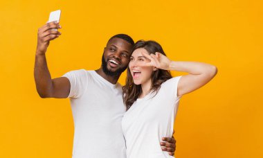 Neşeli ırklar arası çift akıllı telefonlarıyla selfie çekiyor, sarı arka planda birlikte eğleniyorlar. Neşeli kız barış jesti yapıyor, boş alan