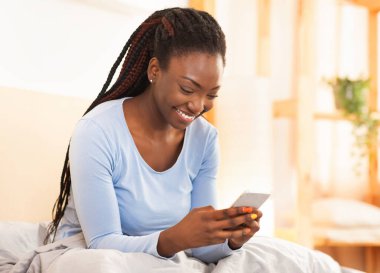 Cep telefonu kullanan neşeli afro kadın evde yatağında oturmuş cep telefonu oyunları oynuyor. Seçici Odaklanma