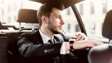 Takım elbiseli, yakışıklı, sakallı genç bir işadamı lüks bir arabada oturuyor ve pencereden şehre bakıyor.