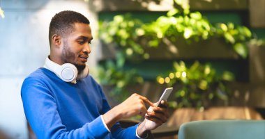 Teknoloji Konsepti. Kulaklıklı genç afro öğrencinin modern kafede oturan akıllı telefonu kullanışının yan görüntüsü