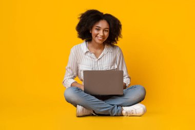 Serbest Kavram. Yerde dizüstü bilgisayarla oturan mutlu siyah kız internette çalışıyor, kopyalama alanı olan sarı stüdyo arka planı