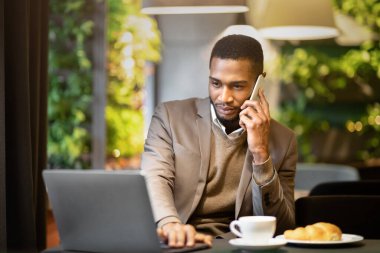 Kurumsal İletişim. Kendine güvenen siyah işadamı cep telefonuyla konuşuyor, laptoptan raporları okuyor.