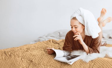 Relaxed blonde girl covered in towel and bathrobe reading magazine, laying on bed at home