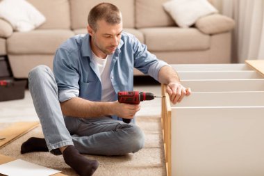 Evde, yerde oturan elektrikli matkap aletini kullanarak rafları birleştiren olgun bir adam. Erkek Ev İşi ve Mobilya Evi. Tamirat sırasında ahşap dolabı tamir eden tamirci.