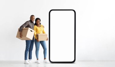 Neşeli siyah bir çift bir arada duruyor, ellerinde karton kutular. Büyük boş bir telefon çerçevesinin yanında konumlanmışlar, neşeli bir atmosfer yaratıyorlar. Gülüşleri heyecan ve takım çalışmasını yansıtıyor..