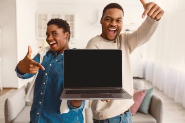 Şu siteyi kontrol et. Heyecanlı Afro-Amerikalı çift ellerinde siyah boş ekranla dizüstü bilgisayarlarını tutuyor ve gösteriyorlar, cihaza boş bir alan sunuyorlar, arka planda bulanıklık var.
