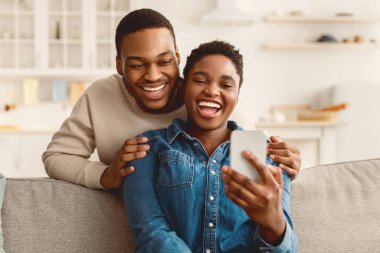Gülümseyen Afro-Amerikan bir adam akıllı telefona bakarken kadınını arkadan kucaklıyor. Sevgili çift sosyal medyayı cep telefonuyla paylaşıyor, mutlu kadın koltukta oturuyor, aletleri gösteriyor.