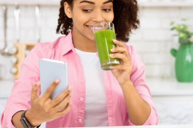 Günaydın Smoothie. Afrikalı bir kadının telefon kullanıp mutfakta ev yapımı detoks suyu içerken çekilmiş yakın plan portresi.