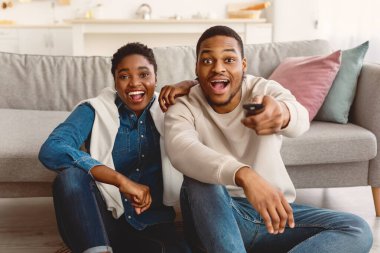 Heyecanlı Afrikalı siyahi çift TV şovu ya da film izliyor, ellerinde uzaktan konsolla evde yerde oturuyor ve oturma odasındaki kanepeye yaslanıyor, kameraya bakan şaşkın adam ve kadın.