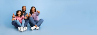 Üç kişilik gülümseyen Afro-Amerikan ailesi, iki eliyle baş parmak işareti yapıyor, iyi bir şeyi onaylıyor ya da öneriyor, mavi stüdyo arka planında, panorama, bedava kopya alanı, yerde oturuyor.