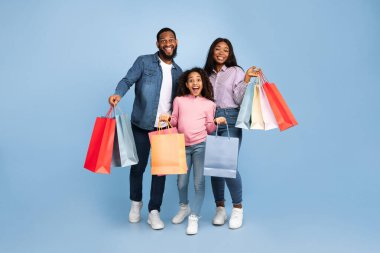 Alışveriş eğlencesi. Afro-Amerikalı neşeli adamın vücut portresi, bayan ve heyecanlı genç kız kağıt torbalarıyla poz veriyor, mavi stüdyoda yan yana duruyor, satışların ve alışverişlerin keyfini çıkarıyor.