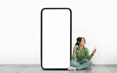 Bir kadın yerde oturuyor, elinde bir kredi kartı ve tablet tutarken gülümsüyor, büyük boş bir akıllı telefon ekranının yanında. Ayarlar rahat bir dijital alışveriş deneyimi olduğunu gösteriyor..