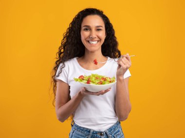 Sağlıklı yemek. Mutlu Latin Gündelik Kadın Portresi Lezzetli Taze Sebze Salatası, Tabak Tabağı ve Çatalla Kameraya Bakıyor. Mutlu Milenyum Kadını Sarı Turuncu Arkaplanda İzole Edildi