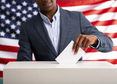 ABD seçimleri 2020. Mutlu Afro-Amerikalı adam, Amerikan bayrağı arka planında oy sandığına kağıt sokuyor.