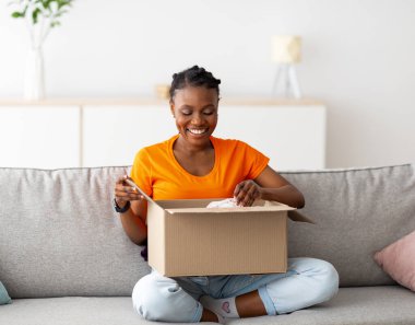 Tam boy, karton paketi açan pozitif siyahi kadın, eve çevrimiçi teslimat alıyor. Genç bayan müşteri, müşteri hizmetlerinden memnun, web sitesi siparişini açıyor.
