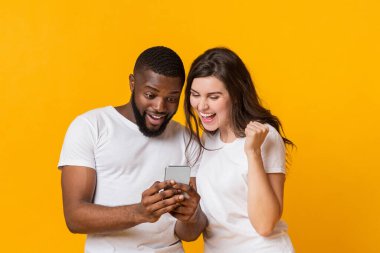 Gezici ikramiye. Şaşırmış çok ırklı çift akıllı telefon ekranına bakıyor ve sevinçle haykırıyor, sarı arka planda birlikte poz veriyorlar,