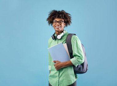 Sırt çantalı ve defterleri kameraya gülümseyen, mavi stüdyo arka planında poz veren zeki Afro-Amerikan öğrenci. Eğitim, üniversite, üniversite, öğrenim, ders kavramı