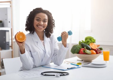 Bağışıklık için spor ve vitamin. Klinikte portakal ve halter tutan mutlu zenci kadın doktor, iş yerinde poz veriyor, boş yer var.