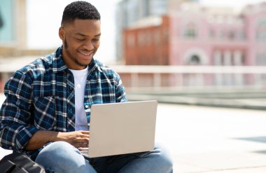 Başarılı bir Freelancer. Mutlu Afrikalı Adam gülümsüyor ve bilgisayarla çalışıyor, halka açık bir parkta oturuyor.
