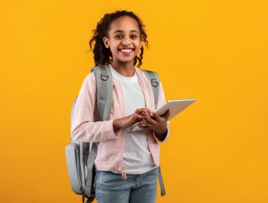Online Eğitim Konsepti. Sırt çantalı mutlu genç öğrencinin portresi e-öğrenim tableti kullanıyor, sarı turuncu stüdyo geçmişi var. Kameraya poz veren mutlu Afrikalı öğrenci