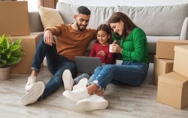 Yeni evlerine taşındıktan sonra dizüstü bilgisayar kullanan üç kişilik genç Müslüman bir aile. Gayrimenkul, Ev Sahipliği ve Taşınma Konsepti. Ebeveynler İnternete Bakıyor, Kız Ekranı Gösteriyor