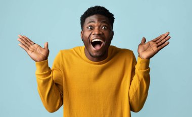 Şaşırmış Afro-Amerikan tarzı, sarı elbiseli genç adam kameraya bakıyor, ağzı açık ve avuçlarını kaldırıyor, mavi stüdyo arka planında hareketler yapıyor, hayret verici, yakın plan çekimler yapıyor.