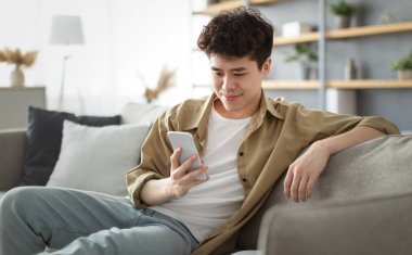 Enjoying Weekend. Handsome Relaxed Asian Man Using Smartphone While Sitting Leaning On Couch At Home, Happy Guy Resting In Living Room, Browsing Internet Or Messaging With Friends, Selective Focus