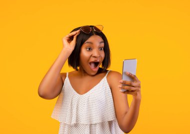 Yazlık kıyafetli siyahi kadın heyecanla akıllı telefona bakıyor turuncu stüdyo arka planında güneş gözlüğünü kaldırıyor. Şaşırmış Afrikalı Amerikalı kadın internette yaz indirimini öğreniyor.